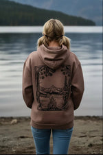Woman in nature hoodie with mountain lake print, standing by water, snow-capped peaks background