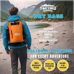 Man wearing orange Tactiko Survival dry bag backpack near mountain lake, ideal for camping, travel, and outdoor adventures.