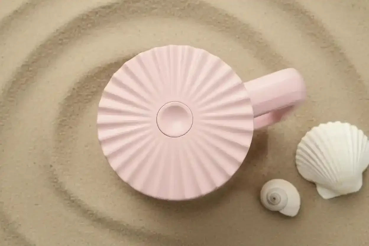 Pink ceramic mug with sunburst design on sandy beach, seashells beside it