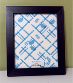 Natural wooden picture frame with Zuri & Zori branding, showing cover protectors, against a green wall.