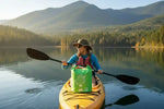 Woman kayaking on calm lake with forested mountains, wearing hat and life vest, green dry bag on yellow kayak