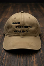 Beige cap with 'Rock Strength Healing' text on a wood floor