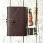 Handmade leather journal with stitching tools on rustic wood background
