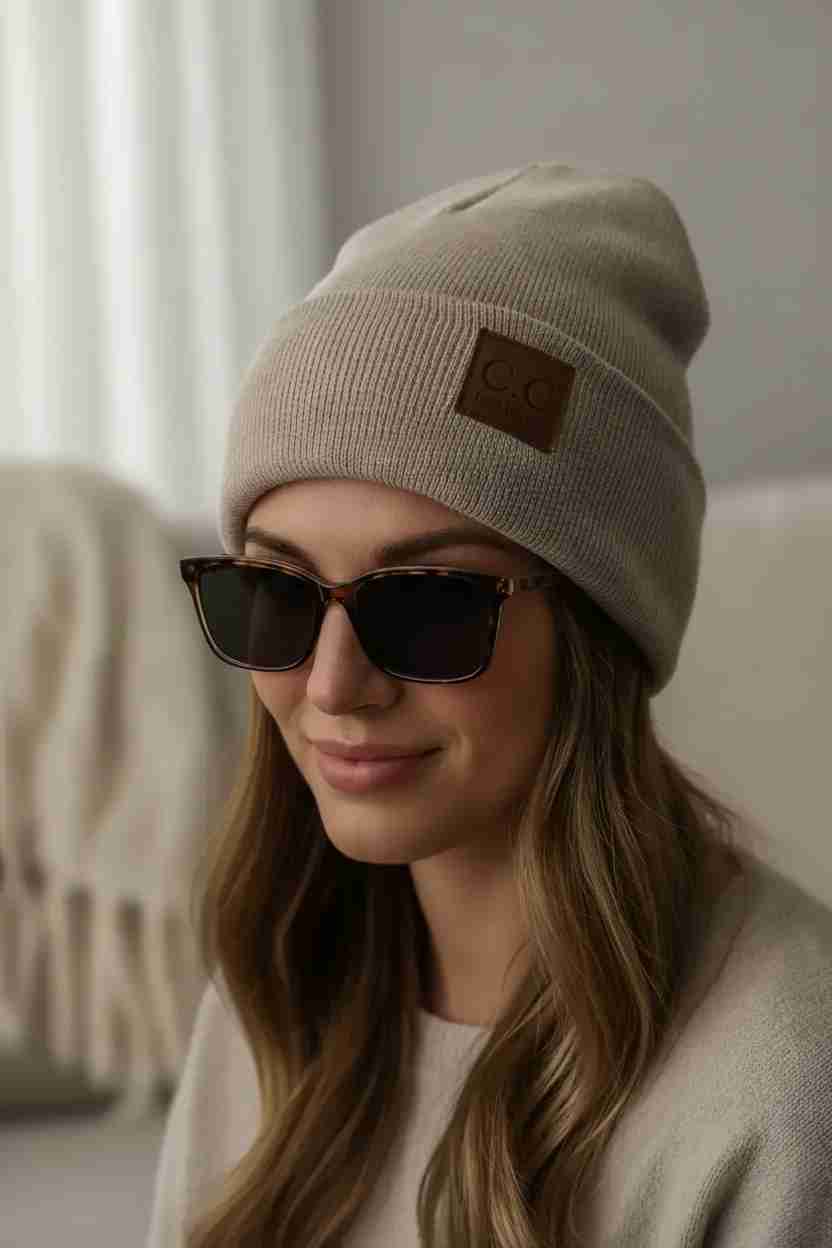 Woman wearing a beige beanie with a patch and dark sunglasses indoors.