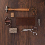 Vintage tools and leather goods on dark wood, including hammer, scissors, stamp, and wallet