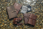 Handcrafted leather journals and notebooks on pebbled shore, water reflecting sunlight