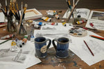  Mugs Two blue ceramic mugs with a visible brand label on an artist’s table Mugs