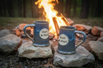 Mugs Two blue ceramic mugs with 'Zuri & Zori' labels on rocks in front of a campfire