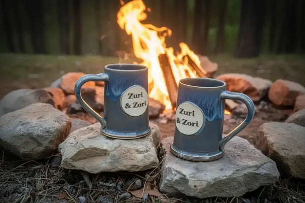 Two blue ceramic mugs with Zuri & Zori logo by a campfire in forest setting