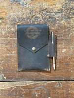 Black leather pen case with snap closure and pen, resting on rustic wooden surface