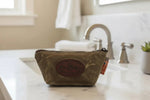 Olive green waxed canvas toiletry bag with leather patch, on marble bathroom counter near sink