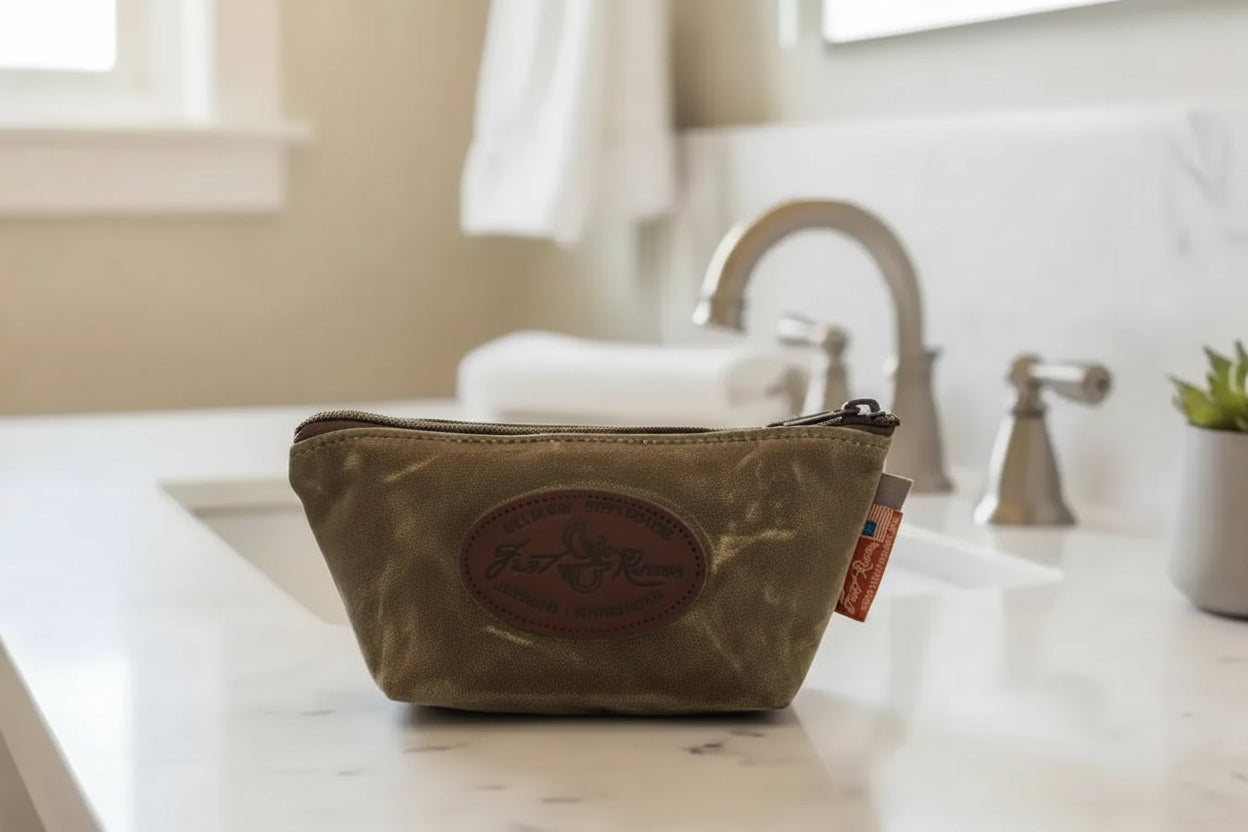 Olive green waxed canvas toiletry bag with leather patch, on marble bathroom counter near sink