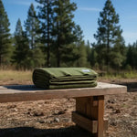 Olive green folded blanket on rustic wooden bench in forest setting under blue sky